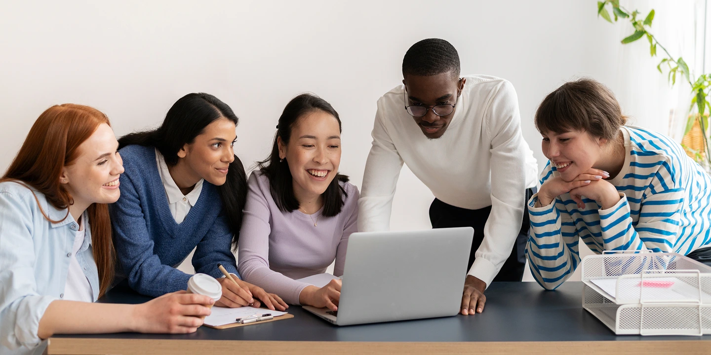 A group of happy students huddled around a desk, focused on a laptop screen.